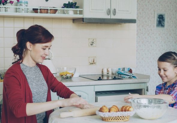 Young mother rolling dough and talking to her little cute daughter while cooking in the kitchen on weekend. Family, food, home and people concept