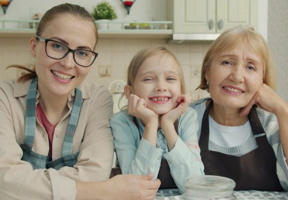 Portrait of joyful family grandmother, mother and daughter smiling in modern kitchen sitting together and looking at camera with happy faces.