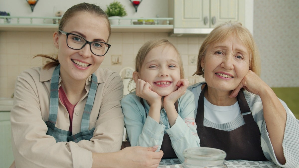 Portrait of joyful family grandmother, mother and daughter smiling in modern kitchen sitting together and looking at camera with happy faces.